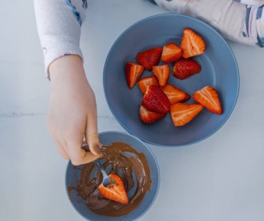 Bir çocuğun taze bir çileği çatalla erimiş çikolataya batırmasının en iyi görüntüsü. Dilimlenmiş çilekli kase. Tatlı hazırlama sahnesi, tatlı ve aile odaklı.