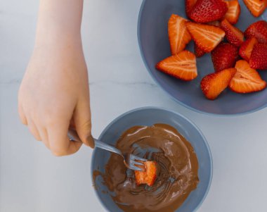 Bir çocuğun elinin taze bir çileği çatalla erimiş çikolataya batırışının en üst görüntüsü. Yanında dilimlenmiş çilek olan bir kase. Tatlı hazırlama, hoşgörü ve yemek zevki kavramı
