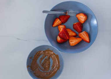 İki mavi kasenin üst görüntüsü: biri erimiş çikolatalı, diğeri dilimlenmiş çilekli ve çatallı. Sıcak yemek tasarımı sahnesi, tatlıya düşkünlüğü ve ev yapımı atıştırmalık anı temsil ediyor.