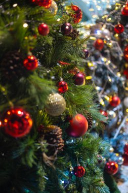 Süslü bir Noel ağacının kırmızı süslemeleri, parlak ışıkları ve çam kozalakları. Bayram havası yumuşak bir odak noktasında bokeh ve sıcak tonlarla yakalanır.