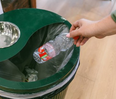 Yakından bakıldığında çöp kutusuna 'Geri Dönüşüm' etiketli boş bir plastik su şişesi atan bir el görülüyor, ekoloji vurgulanıyor, sıfır atık yaşam tarzı ve kapalı bir alanda çevresel koruma
