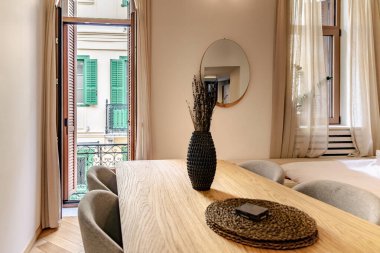 Doğal ahşap ve nötr dekorlu modern bir bej yemek odası. Yeşil panjurlu bir balkona açılan kapı doğal havalandırmayı işaret ediyor.