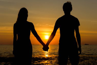 Classical Valentine Day scene with the silhouette of young couple holding hands while contemplating sunset at the beach in the island of Koh Phangan, Thailand. Lovers staring at sun, honeymoon concept