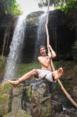 Young man in trunks hanging from vine in rain forest with waterfall on the background in Banlung, Cambodia. Fun adventure traveler concept
