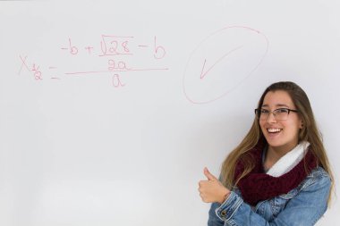 Happy blonde girl with thumb up smiling to correct math equation written on white board. Young woman achieves good marks in college. Perfect student concept