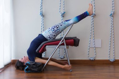 Female yogini using props to prepare back bend posture like urdhva dhanurasana. Iyengar yoga style teacher resting on chair, mats and shoulders on blankets in studio