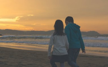 Lovely scene of kids walking together holding hands on empty beach at sunset in Atxabiribil, Basque Country. Little boy and girl at twilight by the sea. Vintage effect