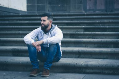 Kilisenin merdivenlerinde oturan düşünceli genç adam. Gri kapüşonlu ve kot pantolonlu şık bir erkek model sokakta tek başına. Kentsel şehirde soğuk mavi tonlar