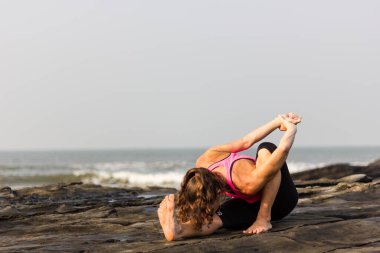 Woman practicing yoga early morning on the rocks by the sea in Goa beach, India. Female yogi on marichyasana pose