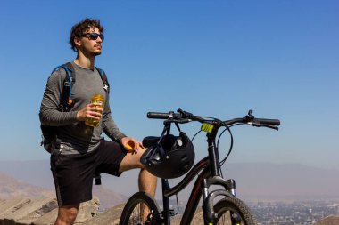 Young biker with sunglasses holding water bottle next to mountain bike on sunny day. Handsome man taking a break while and enjoying splendid views of Santiago city in Chile clipart