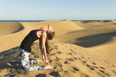 Young woman practices yoga in Maspalomas sand dunes at sunset. Female yogi on camel pose, known as ustrasana, at Gran Canaria desert on sunny day. Sports activity in natural environment clipart