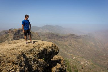 Tourist on mountain edge facing great landscape views. Young man on blue t shirt standing on top of rock on sunny day in Roque Nublo. Adventure, explore, freedom, visionary, solitude concepts