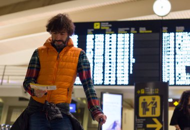 Young man wearing casual winter clothes with carry on luggage looks at boarding pass with flight schedule board on the background at airport. Trasportation, departure, arrival concept