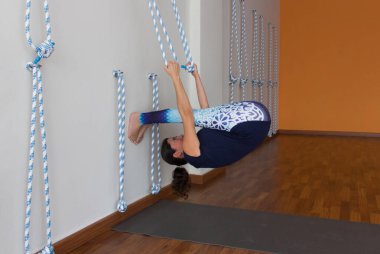 Young yogini doing paschimottanasana upside down grabbing ropes on white wall in studio. Female yoga teacher on reverse forward bend in empty center room. Discipline, self practice concept