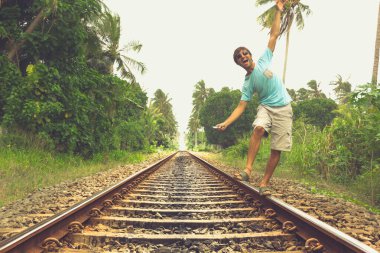 Young man walking on railroad track while listening to music with headphones on his smartphone in rural area in Bentota, Sri Lanka. Vintage effect. Risk, reckless, danger, dare, no fear concepts