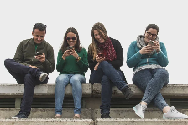 Three young women and one man sitting on grandstands using cellphones. Group of friends laughing together while looking at smartphones. Millennial generation, social media sharing concept
