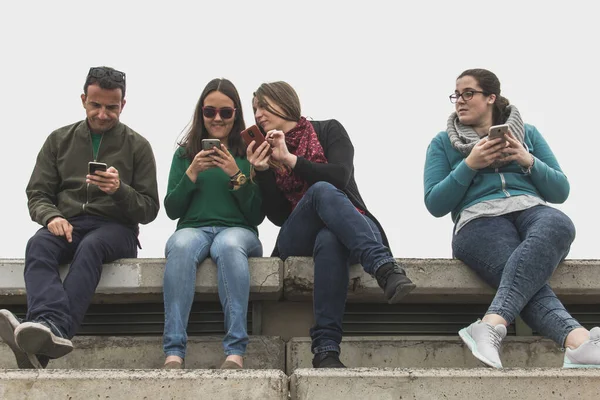 Friends sharing information on cellphones, one girl jealous on the side. Young people using smart phones while sitting on stands with winter clothes. Social media concept