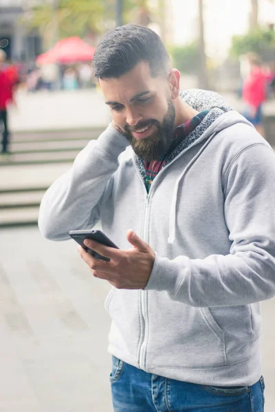 Smiling Young Man Grey Hoodie Holds Cellphone Shy Embarrassed Expression — Φωτογραφία Αρχείου