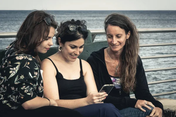 Three young women looking at cellphone sitting on bench by the sea. Telephone overuse, mobile addiction concepts. Vintage effect applied
