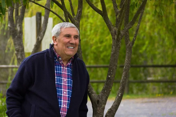 Senior man smiling leaning on tree at park. Retired happy man outdoors with dark blue sweater and colorful checkers shirt. Positive attitude concept