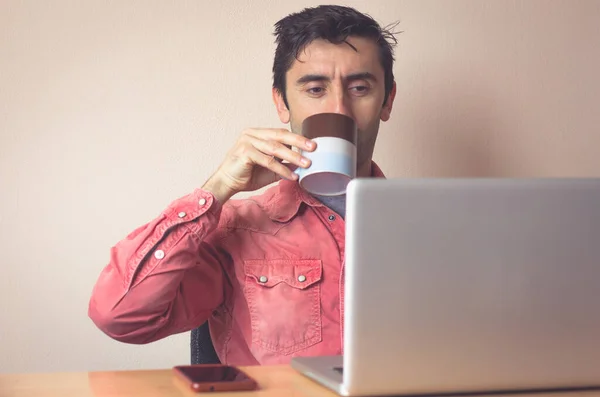 Młody Człowiek Czerwonej Koszuli Pijący Kawę Patrząc Ekran Laptopa Biurku — Zdjęcie stockowe