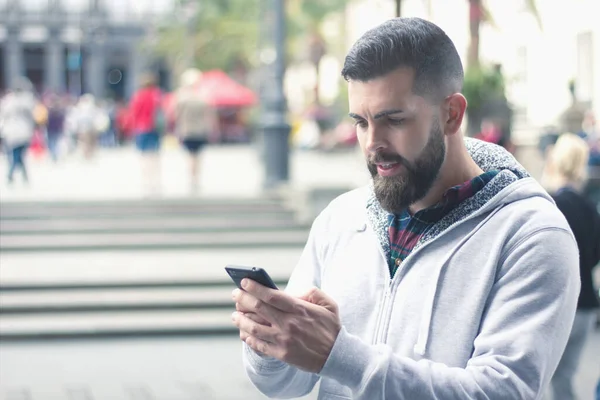 在城市里 满脸胡须的男人在户外用手机 有灰色帽衫的年轻嬉皮士在街上用黑色移动设备发短信 无线因特网概念 — 图库照片