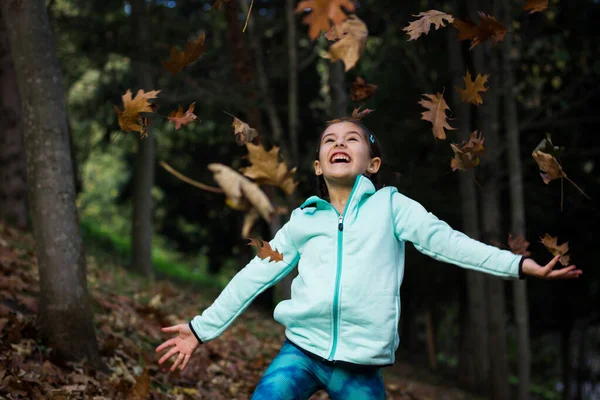 Worry free little girl with open arms looking up at brown leaves in the air in park. Happy kid enjoying autumn season in forest. Child fashion model, freedom, inspiring moment concepts