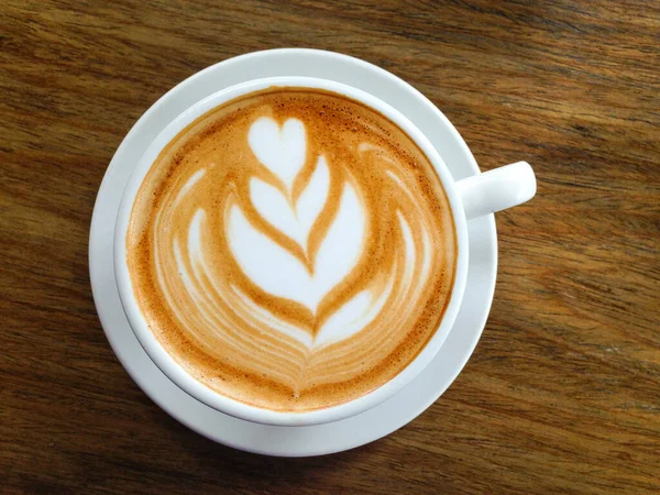 Delicious coffee cappuccino with white milk leaf decoration on white cup and plate over wooden background. Cafe perfect break concept
