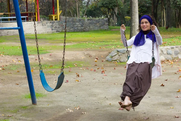 Mutlu, olgun bir kadın, kafasında mor bir şalla sonbahar sezonunda parkta sallanıyor. Çocuk parkında orta yaşlı bir kadın
