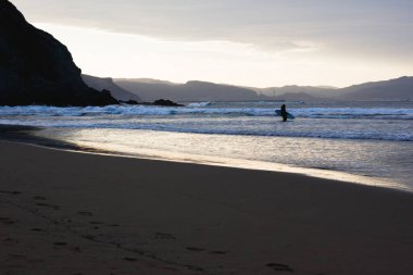 Gün batımında Bask Bölgesi 'ndeki boş Atxabiribil plajında sörf tahtasıyla suya giren kadın silueti. Kışın sörf yapma tutkusu