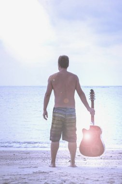 Young man holding his guitar while standing up looking out to the sea in the island of Koh Phangan, Thailand. Musician looking for inspiration in nature. Lens flare and blue filter applied clipart