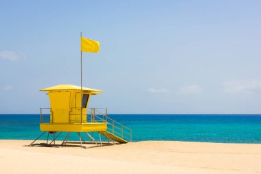 Fuerteventura Corralejo doğal parkındaki boş plajda görevli renkli cankurtaran istasyonu. Turkuaz denizde küçük bir balıkçı teknesi. Güneşli bir günde, sahil gözlem kulesinin tepesinde sarı bayrak var.