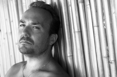 Tayland 'ın Koh Phangan adasındaki plajda bambu kamışlara yaslanan düşünceli görünümlü siyah beyaz genç adam portresi. Sakallı bir erkek model.