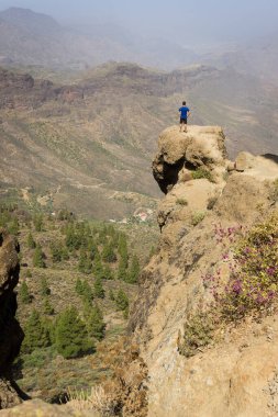 Güneşli bir günde Roque Nublo, Gran Canaria 'da, kayanın tepesinde tek başına duran mavi gömlekli adam. Dağ kenarındaki kaşif panoramik manzaraya bakıyor. Açık hava aktivitesi, macera, baş dönmesi, özgürlük kavramları