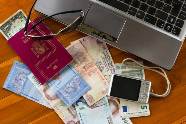 Silver laptop by a digital camera, sunglasses, note bills from different countries and US dollars inside a Spanish passport on wood table, top perspective. Translation: European Union, Spain, passport