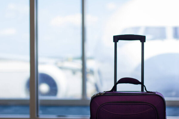 Purple carry on luggage standing by window of boarding gate at airport with airplane on the background