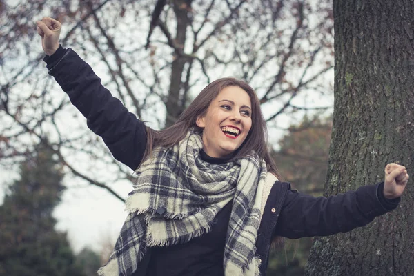 Joyful brunette girl smiling with arms up in the park on cloudy autumn season day. Celebration concept. Pretty young woman happy as if having won the lottery
