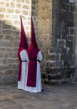 Kırmızı beyaz giyinmiş iki kişi taş bir duvarın önünde duruyor.