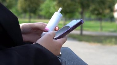 Kadınların elleri cep telefonunda mesaj yazıyor. Sonbahar parkında bir kadının ellerine yakın çekim. Cep telefonu ekranında yazan ve elektronik sigara içen bir kadın.