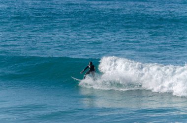 Fotoğrafta arka planda mavi deniz olan bir dalgada sörf yapan bir adam var.