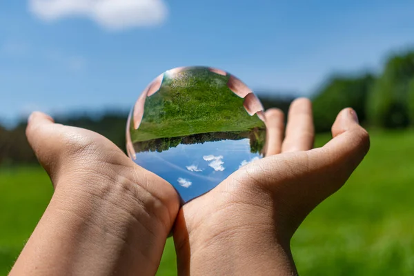 In the photograph you can see the hands of a child holding a crystal ball where you can see a green meadow and everything that surrounds it out of focus.