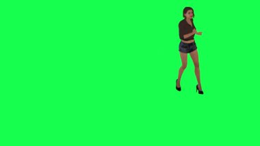 Kahverengi elbiseli, yüksek topuklu ve siyah şortlu genç bir kadın yeşil ekranın önünde daireler çiziyor ve egzersiz yapıyor.