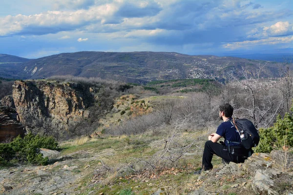 stock image A man with dark clothes and a backpack sitting and staring to Momini scali (Momina skala) near the villages of Hrabrino and Izvor in Rhodope Mountains, Bulgaria. A beautiful panorama on a sunny day in early spring.