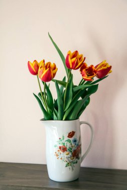 Tahta bir rafta kırmızı ve sarı lale buketi içeren çiçek desenli beyaz bir porselen sürahi. Seçici odak ve kopyalama alanı olan dikey resim