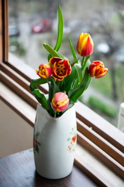 Pencerenin yanında kırmızı ve sarı laleler bulunan beyaz bir porselen sürahi. Seçici odak ve bulanık arkaplan ile dikey resim