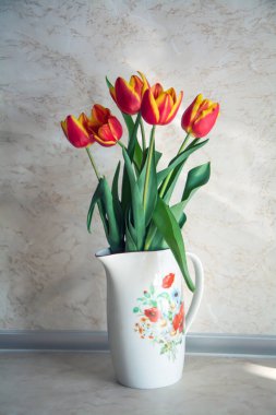 Mutfak tezgahında kırmızı ve sarı lale buketi bulunan çiçek desenli beyaz bir porselen sürahi. Seçici odaklı dikey resim.