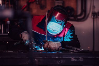 Profesional welder in protective uniform and mask welding metal pipe on the industrial table with other workers behind in the industrial workshop