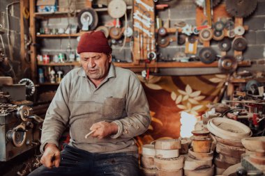 A senior man sitting in the workshop and processing wooden utensils in the old manual way. 