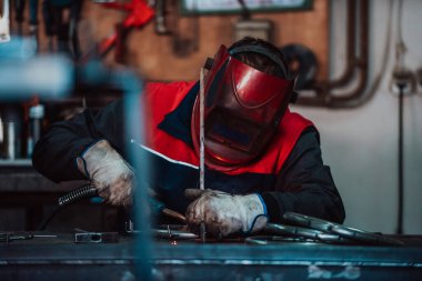 Profesional welder in protective uniform and mask welding metal pipe on the industrial table with other workers behind in the industrial workshop
