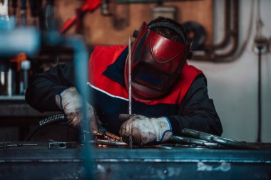 Profesional welder in protective uniform and mask welding metal pipe on the industrial table with other workers behind in the industrial workshop